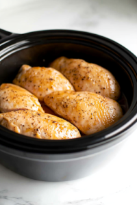 A black slow cooker on a white marble cooktop, with four boneless, skinless chicken breasts neatly placed inside. The chicken is evenly arranged, ready to absorb the rich barbecue flavors during slow cooking.