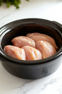 A black slow cooker on a white marble cooktop, with six raw chicken breasts neatly arranged at the bottom, ready for slow cooking to create a flavorful Italian-inspired dish.