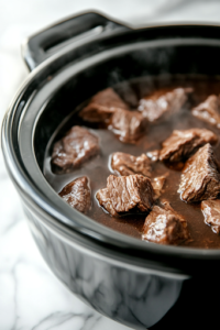 A black slow cooker on a white marble cooktop, now covered and simmering, as the beef slowly cooks for hours, absorbing the aromatic broth and becoming tender.