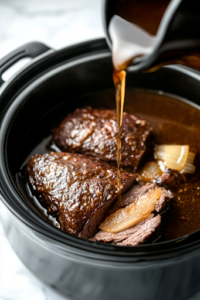 A black slow cooker on a white marble cooktop, as a rich blend of beer, beef broth, and French onion soup is poured over the rump roast, filling the pot with deep, savory flavors.