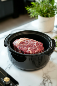 A black slow cooker sitting on a white marble cooktop, with a raw 4-pound rump roast placed inside, ready for slow cooking to achieve a tender and flavorful French Dip filling.
