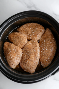 A black slow cooker on a white marble cooktop, with four boneless, skinless chicken breasts neatly arranged and evenly coated with homemade Italian seasoning, onion powder, kosher salt, and cracked black pepper, ready for slow cooking.