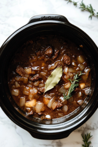 A close-up shot of a black slow cooker on a white marble countertop, filled with deeply caramelized onions. A steady stream of cooking sherry is being poured in, followed by rich beef broth, fresh thyme sprigs, and a single bay leaf floating on top. The ingredients come together to create a flavorful, aromatic base for the soup.