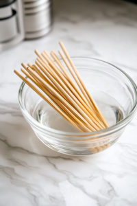 A glass bowl filled with water on a white marble cooktop, with wooden skewers fully submerged to prevent burning during grilling. The water glistens under the light, ensuring the skewers are well-soaked.