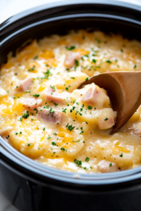 A close-up shot of the black slow cooker on the white marble cooktop, with a wooden spoon gently stirring the creamy scalloped potatoes. The ham and cheese are evenly mixed throughout, ensuring every bite is packed with flavor.
