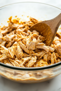 A wooden spoon stirs the seasoned shredded chicken mixture in a glass mixing bowl on the white marble cooktop. The curry powder blends into the mayo, creating a smooth and golden coating.