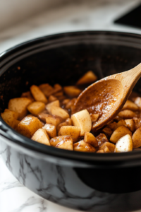 A close-up shot of a wooden spoon gently stirring the softened apples inside the black slow cooker on the white marble cooktop. The apples are breaking down, releasing sweet and spiced aromas throughout the kitchen.