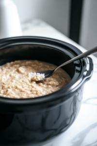 A black slow cooker on the white marble cooktop, with a spoon stirring in salt and half-and-half to make the oatmeal creamy. The smooth consistency is coming together perfectly