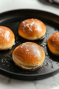 A close-up of a black non-stick pan on the white marble cooktop, where soft hamburger buns are toasting in melted butter. The buns develop a crisp golden-brown crust, creating the perfect base for pulled pork sandwiches.
