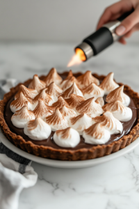 Close-up shot of the fully chilled chocolate pie on the white marble cooktop, topped with fluffy vegan meringue. A hand carefully uses a blow torch to toast the meringue peaks to a golden brown.