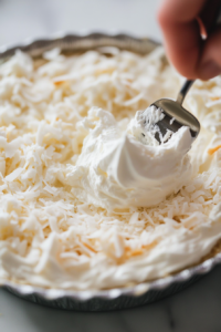 Whipped cream is being spread or piped onto the chilled coconut cream pie, with a smooth, fluffy top. Optional coconut garnish is added on top.