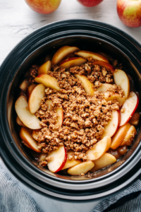 The slow cooker on a wooden countertop, now with the crumbly walnut topping sprinkled evenly over the apples. The crisp is ready to cook.