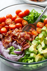 A close-up of a large glass mixing bowl on a white marble cooktop, filled with crisp romaine lettuce, juicy halved cherry tomatoes, thinly sliced red onions, creamy avocado slices, and golden bacon crumbles. A serving spoon gently tosses the fresh ingredients together.