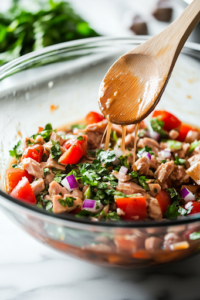 A close-up shot of a large glass mixing bowl on a white marble cooktop, with tuna, diced eggs, chopped celery, pickles, and onion inside. A wooden spoon rests in the bowl, ready to mix the ingredients together.