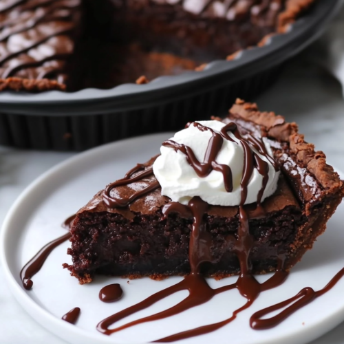 Close-up shot of a sliced piece of vegan brownie pie on a white marble plate, topped with a swirl of vegan whipped cream and drizzled with melted chocolate. The rest of the pie remains in the black pie pan, ready to be served.
