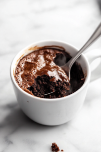 A freshly cooked chocolate mug cake sits on a white marble cooktop, slightly cooled. A spoonful is taken out, revealing its moist and fluffy interior, with gentle steam rising from the warm cake.