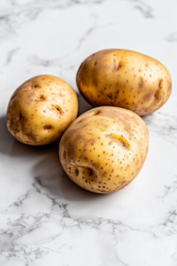 A close-up shot of four baking potatoes resting on a white marble cooktop, freshly washed and scrubbed clean. A soft kitchen towel is placed nearby as the potatoes are gently patted dry, ensuring they are prepped for baking.