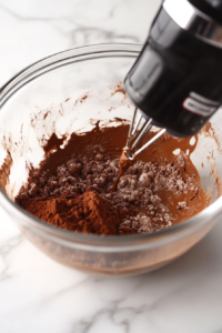 A close-up shot of a mixing bowl where heavy cream, powdered sugar, cocoa powder, and vanilla extract are being whipped until stiff peaks form.