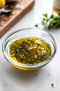 Side view of a small bowl containing olive oil, red wine vinegar, lemon juice, minced garlic, Dijon mustard, oregano, salt, and pepper, being whisked together to form a smooth Greek salad dressing.
