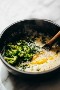 A black saucepan on a white marble cooktop with steamed broccoli and a squeeze of lemon juice being added to the creamy soup. The bright green broccoli contrasts with the smooth, creamy texture.