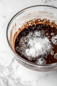 A glass mixing bowl on a white marble cooktop, as sifted confectioners’ sugar and vanilla extract are stirred into the melted chocolate. The rich, dark mixture is thick and velvety.