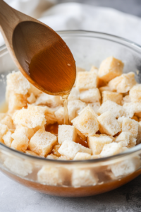 Chicken broth being gradually poured into a glass mixing bowl over the bread mixture, with a wooden spoon gently stirring to moisten the cubes.