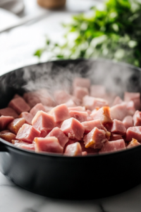 A close-up of diced ham being added to the black Dutch oven on the white marble cooktop. Steam rises as the ham begins to brown, blending with the softened vegetables to enhance the soup’s depth of flavor.