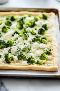 A close-up of the golden-baked pizza crust on a parchment-lined baking sheet, now topped with Parmesan cheese, creamy garlic sauce, fresh broccoli florets, and shredded mozzarella. The toppings are spread evenly, ready for baking.