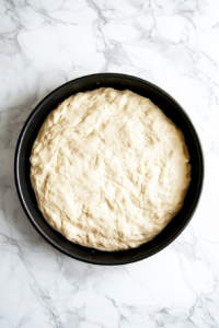 Close-up shot of a 10-inch black pie pan on the white marble cooktop, with an even layer of dough pressed into it. The crust is freshly baked, with a light golden hue forming around the edges.