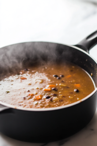 A black skillet on a white marble cooktop as creamy coconut milk, vegetable broth, and sour cream (if using) are poured into the sautéed vegetables. A wooden spoon stirs the rich, golden mixture.