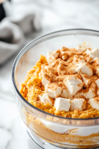 Close-up shot of a glass food processor on the white marble cooktop, filled with smooth organic pumpkin purée, maple syrup, arrowroot powder, pumpkin pie spice, almond butter, tofu, and vanilla extract. The mixture is being blended into a creamy, vibrant orange filling.