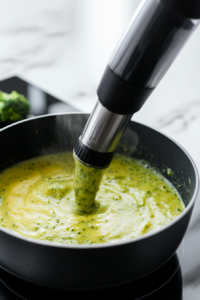 n immersion blender blending the soup in a black saucepan on a white marble cooktop. The soup turns smooth and creamy, with vibrant vegetables swirling into a velvety texture.