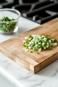 A close-up of a wooden cutting board on a white marble cooktop, where a sharp knife slices through bright red Roma tomatoes. Finely chopped scallions sit neatly in a small glass bowl, prepped and ready for garnishing the tacos.