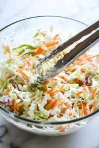 A glass mixing bowl on the white marble cooktop contains a fresh coleslaw mix. A drizzle of creamy dressing made from Greek yogurt, lime juice, olive oil, salt, and pepper is being poured over the slaw. A pair of tongs rests on the side, ready for tossing.