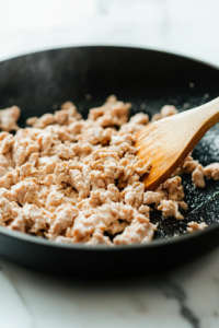 A close-up of a black skillet on a white marble cooktop, now filled with ground chicken breast. The chicken is breaking apart with a wooden spatula as it turns from pink to golden brown, cooking evenly in the pan.