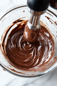 A glass mixing bowl on a white marble cooktop, with an electric hand mixer whipping heavy cream into soft peaks. The creamy, airy texture is forming, ready to be folded into the chocolate.