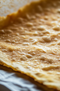 A close-up of the pizza crust baking in the oven, turning golden brown with a slightly crispy texture. The edges are slightly puffed, and the parchment paper remains underneath for support.