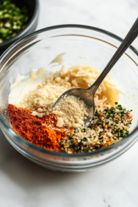 A glass bowl filled with mayonnaise, grated Parmesan cheese, Ranch seasoning mix, garlic powder, and onion powder sits on the white marble cooktop. A spoon is partially submerged in the mixture, ready to combine the ingredients into a creamy coating.