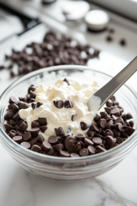 A glass mixing bowl on a white marble cooktop, filled with semisweet chocolate chips and heavy cream. A spatula stirs the melted chocolate mixture, which is smooth and glossy after being heated.