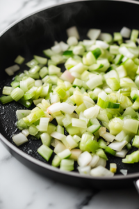 Diced onions and sliced celery sizzling in a black skillet on a white marble cooktop, turning soft and translucent
