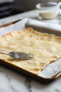 A close-up of the rolled-out pizza crust on a parchment-lined baking sheet over a white marble cooktop. A fork gently pokes holes into the dough to prevent bubbles from forming during baking.