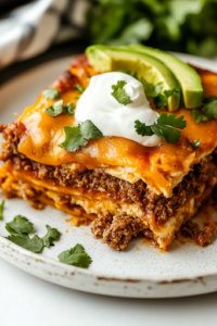 A close-up of a sliced portion of taco casserole on a plate, garnished with fresh cilantro, sour cream, and a slice of avocado. The layers of seasoned beef, creamy eggs, and melted cheese are visible, making each bite look hearty and delicious.