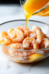 Freshly squeezed lime, lemon, and orange juice is being poured into a glass mixing bowl filled with raw shrimp. The citrus juices coat the shrimp, initiating the curing process on a white marble cooktop