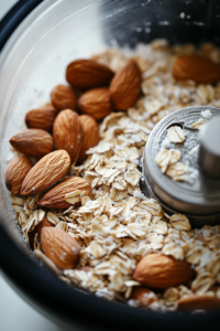 A food processor blending oats, almonds, salt, and sugar into a fine meal. Melted coconut oil is being drizzled in as the mixture turns into a dough-like consistency.