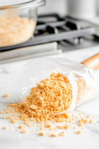A ziplock bag filled with Ritz crackers rests on the white marble cooktop, being crushed by a rolling pin. The golden crumbs start to break apart, spreading slightly around the bag, getting ready for the crispy coating.