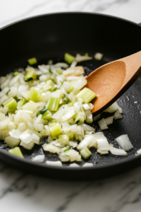 A black skillet on a white marble cooktop, with a pat of butter melting. Chopped onions and celery sizzle in the pan, turning soft and translucent, as a wooden spoon gently stirs them.