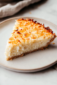 Close-up shot of a slice of coconut custard pie on a plate, with a creamy, golden custard texture. The slice sits on the white marble cooktop with a fork ready to take a bite.