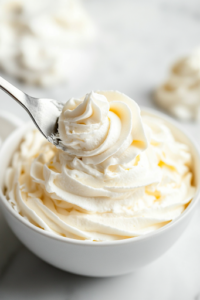 A bowl filled with perfectly whipped French buttercream sits on a white marble cooktop. A spatula lifts a smooth, silky swirl, showing its luscious texture, ready for frosting.