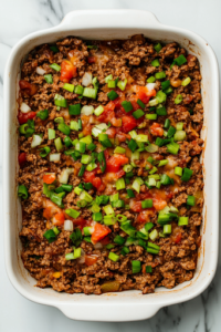 A greased baking dish with the seasoned taco beef mixture evenly spread across the bottom. The bright colors of green onions and salsa contrast beautifully with the deep brown beef, making it look flavorful and ready for the next step.