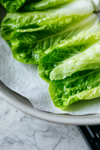 A close-up of fresh romaine lettuce leaves being washed under running water over the white marble cooktop. A paper towel gently pats them dry, ensuring they remain crisp and fresh for taco assembly.